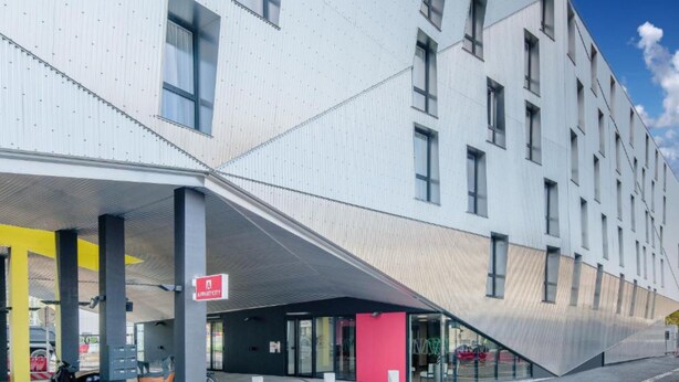 A modern building with a unique angular design, featuring a covered entrance area with a bike rack and a sign indicating a public space.