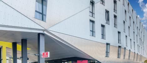 Un edificio moderno con un diseño angular distintivo, que cuenta con un acceso cubierto, un aparcamiento para bicicletas y un letrero que indica un espacio público.