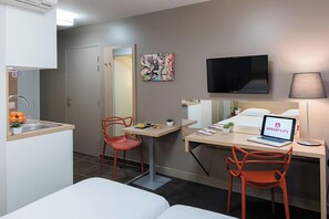 Een hotelkamer met een bed, een bureau met een laptop, een televisie, een stoel, een klein tafeltje met fruit en een wastafel.