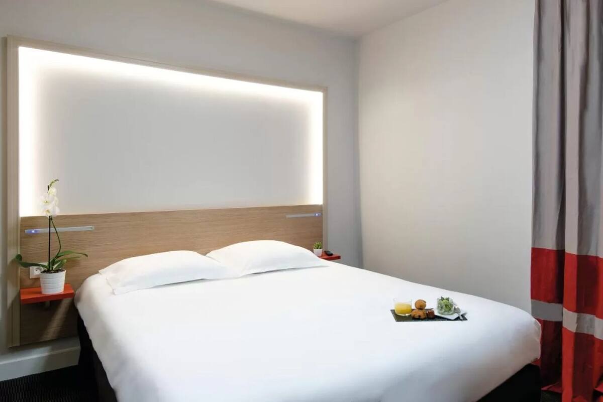 Een hotelkamer met een opgemaakt bed, een nachtkastje met een plant en een rood-wit gestreept gordijn.