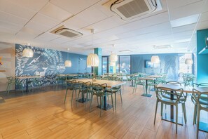 Een ruime eetruimte met meerdere tafels en stoelen, een hoge plafond met ventilatieopeningen en een muurschildering.