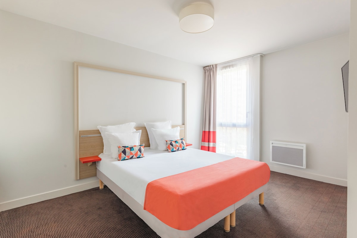 Una habitación de hotel con cama, mesita de noche, ventana con cortinas y un calentador de pared.