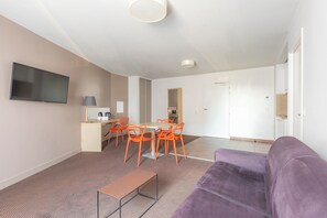 Een moderne woonkamer met een bank, een eettafel met stoelen, een flatscreen televisie aan de muur en een keuken in de achtergrond.