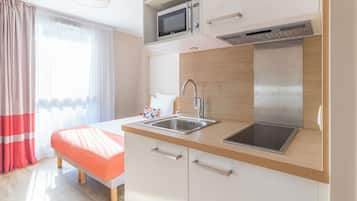 Une cuisine moderne équipée d’un four à micro-ondes, d’un évier et d’une cuisinière, un lit recouvert d’une couverture rouge et une fenêtre ornée de rideaux légers.
