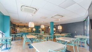 A spacious restaurant with teal chairs and tables, a bar area, and pendant lighting.