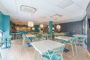 A spacious restaurant with teal chairs and tables, a bar area, and pendant lighting.