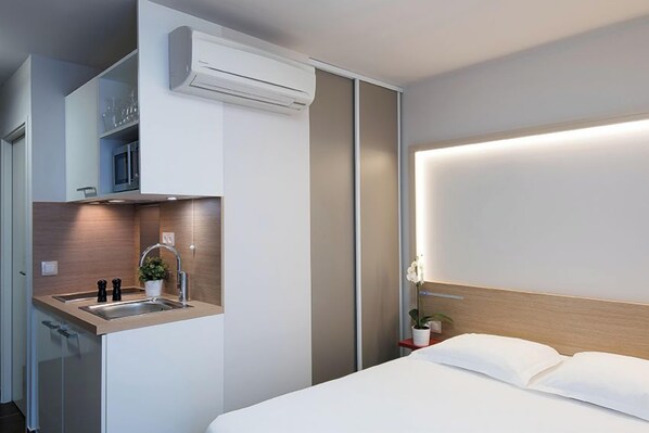 A modern hotel room with a bed, a bedside table with a plant, a built-in sink, and a microwave.