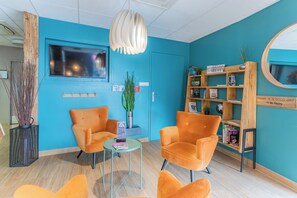A reception area with two orange chairs, a small table, a bookshelf, and a television on the wall.