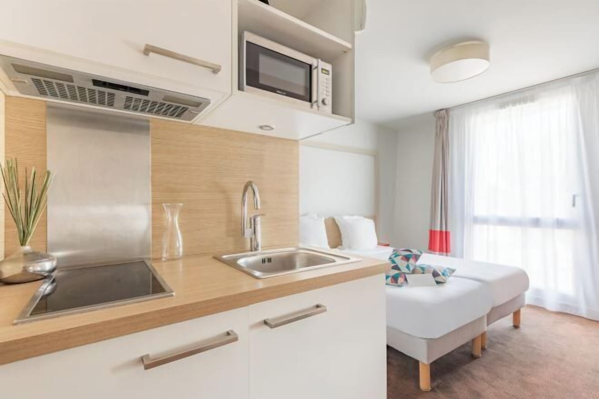 A modern hotel room with a kitchenette, a bed, a window with curtains, and a ceiling light.