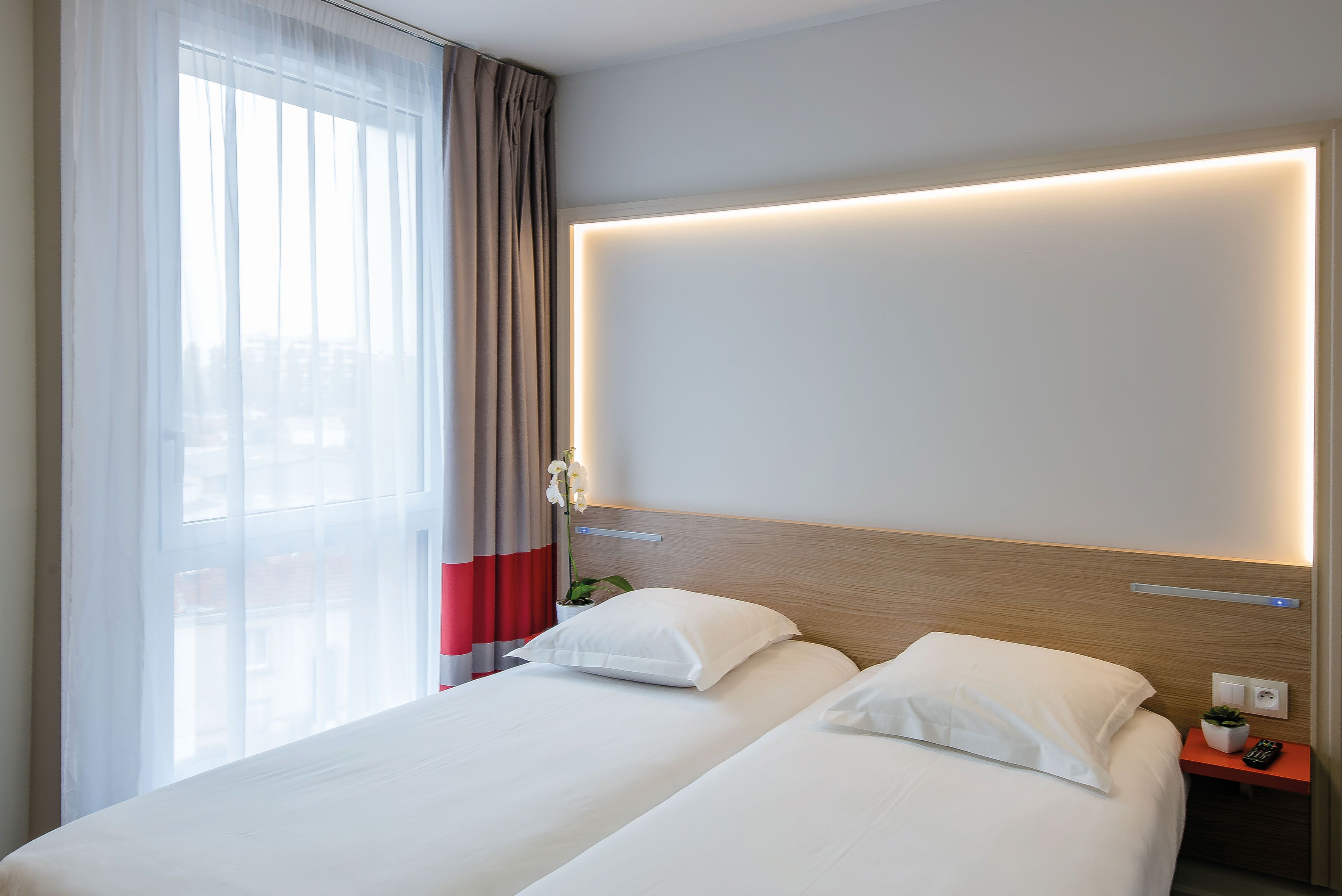 Habitación de hotel con dos camas, una mesita de noche con un control remoto, una lámpara de pared y un ventanal con cortinas traslúcidas.