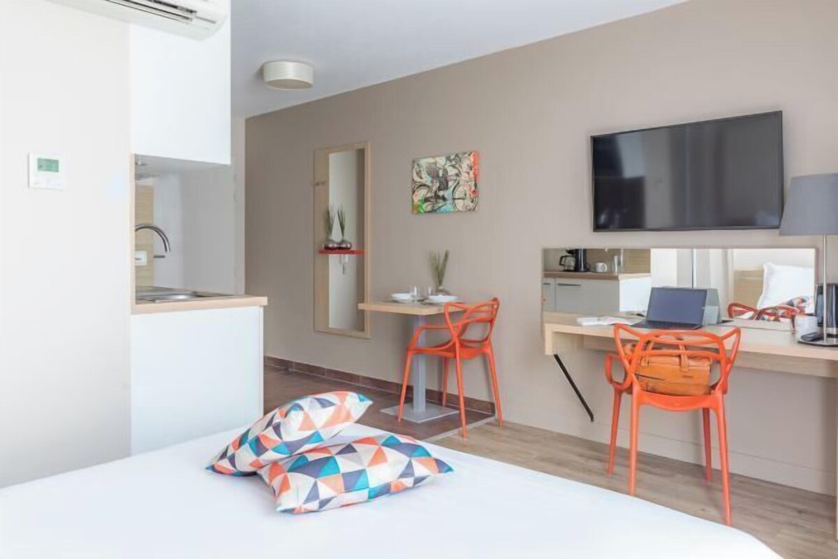 Una habitación de hotel moderna con cama, escritorio, computadora portátil, televisor y pequeña cocina.
