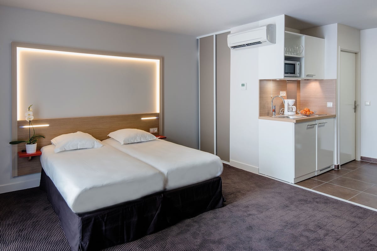 Une chambre d’hôtel comprenant un lit, des tables de chevet, une petite plante en pot, un climatiseur fixé au mur et un coin cuisine.