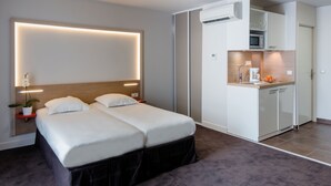 A hotel room with a bed, bedside tables, a small potted plant, a wall-mounted air conditioning unit, and a kitchenette area.