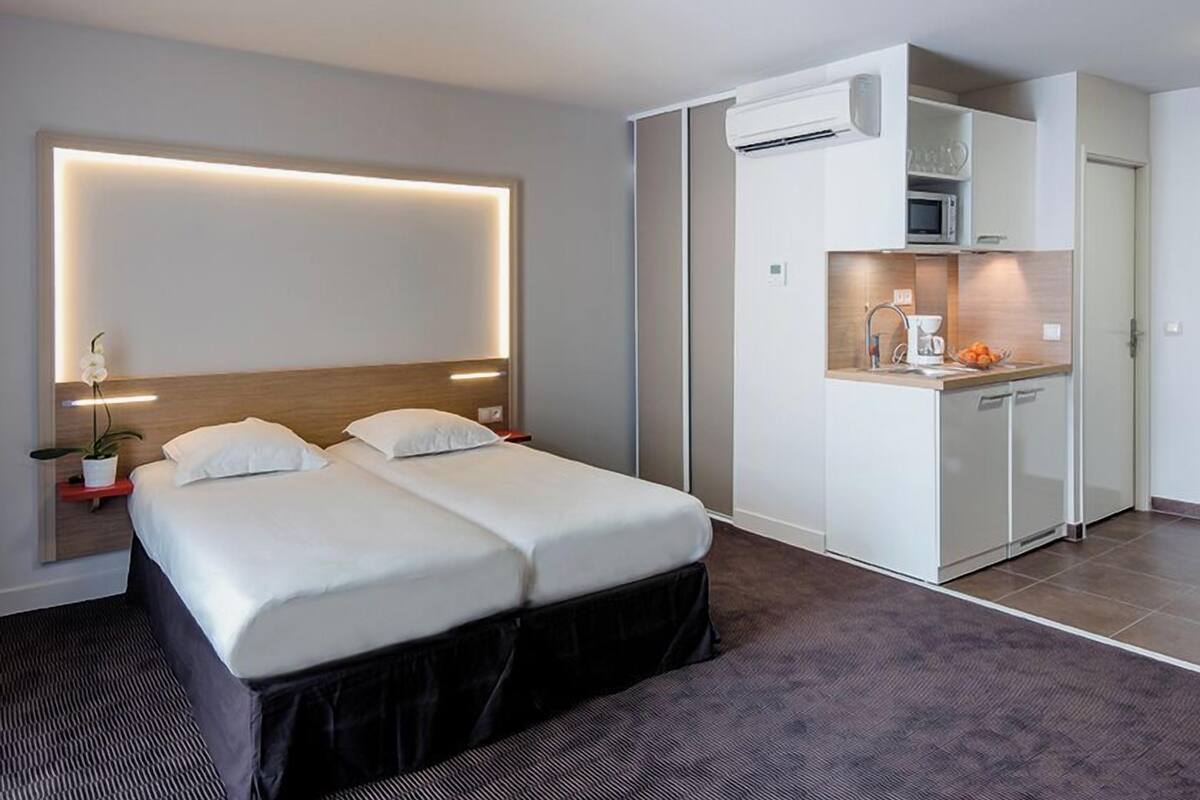Een moderne hotelkamer met een groot bed, een kleine kitchenette en een nachtkastje met een lamp.