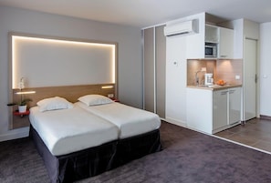 A hotel room with a bed, bedside tables, a small kitchen area, and an air conditioning unit.