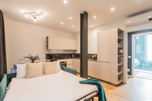 A modern hotel room with a bed, bedside table, and a view of the kitchen and balcony.