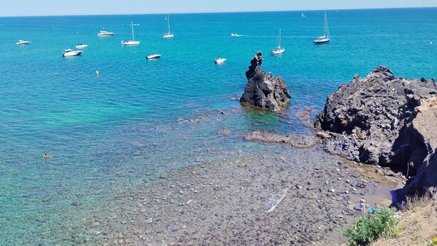 A rocky shoreline with clear turquoise water, boats, and a prominent rock formation.