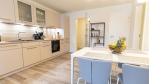 A modern kitchen with light wood cabinetry, a white countertop, and a dining area with a white table and blue chairs. - Anchorage 3 - Terrace. Sauna. Dogs allowed. Barrier-free & close to the beach. (Kellenhusen)