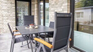 A modern outdoor dining area with a brick wall, glass doors, and a view of a residential area. - Anchorage 3 - Terrace. Sauna. Dogs allowed. Barrier-free & close to the beach. (Kellenhusen)