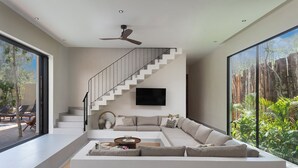 Ein modernes Wohnzimmer mit einem großzügigen Ecksofa, einem flachbildfernseher an der Wand, einer Treppe mit Glastrennwand und Blick ins Grüne durch große Fenster.