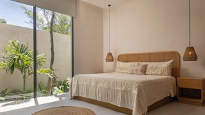 Ein modernes Schlafzimmer mit einem großen Bett, zwei Nachttischlampen, einem Holznachttisch und Blick auf Grün hinter einer Glastür.