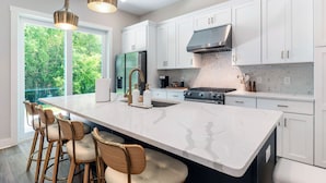 A modern kitchen with white cabinets, a large island with bar stools, stainless steel appliances, and a view of greenery outside. - 3Zs Beach House 30A  Sleeps 10! (Inlet Beach)