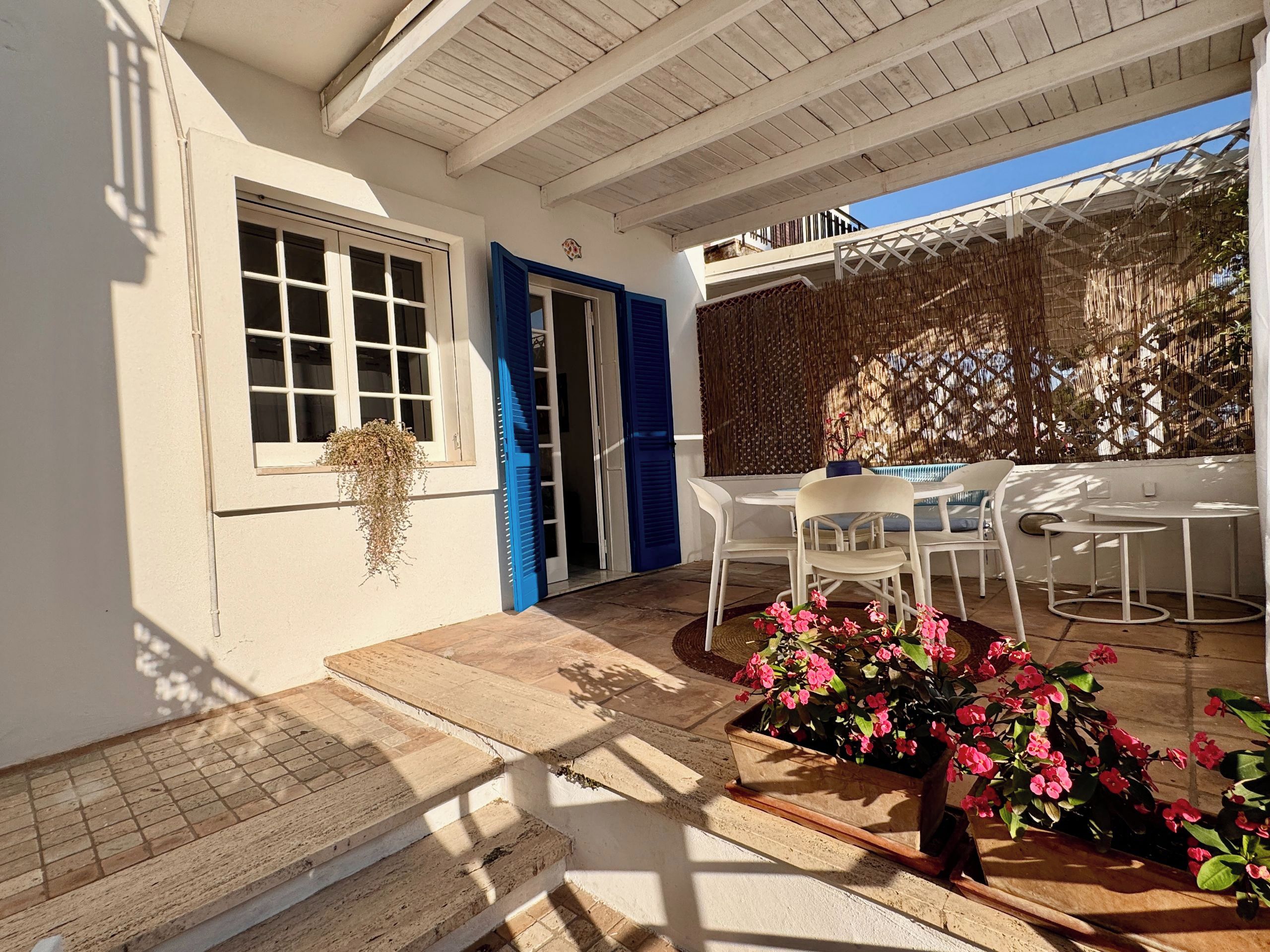 A covered outdoor seating area with white tables and chairs, a blue door, and a window with shutters.
