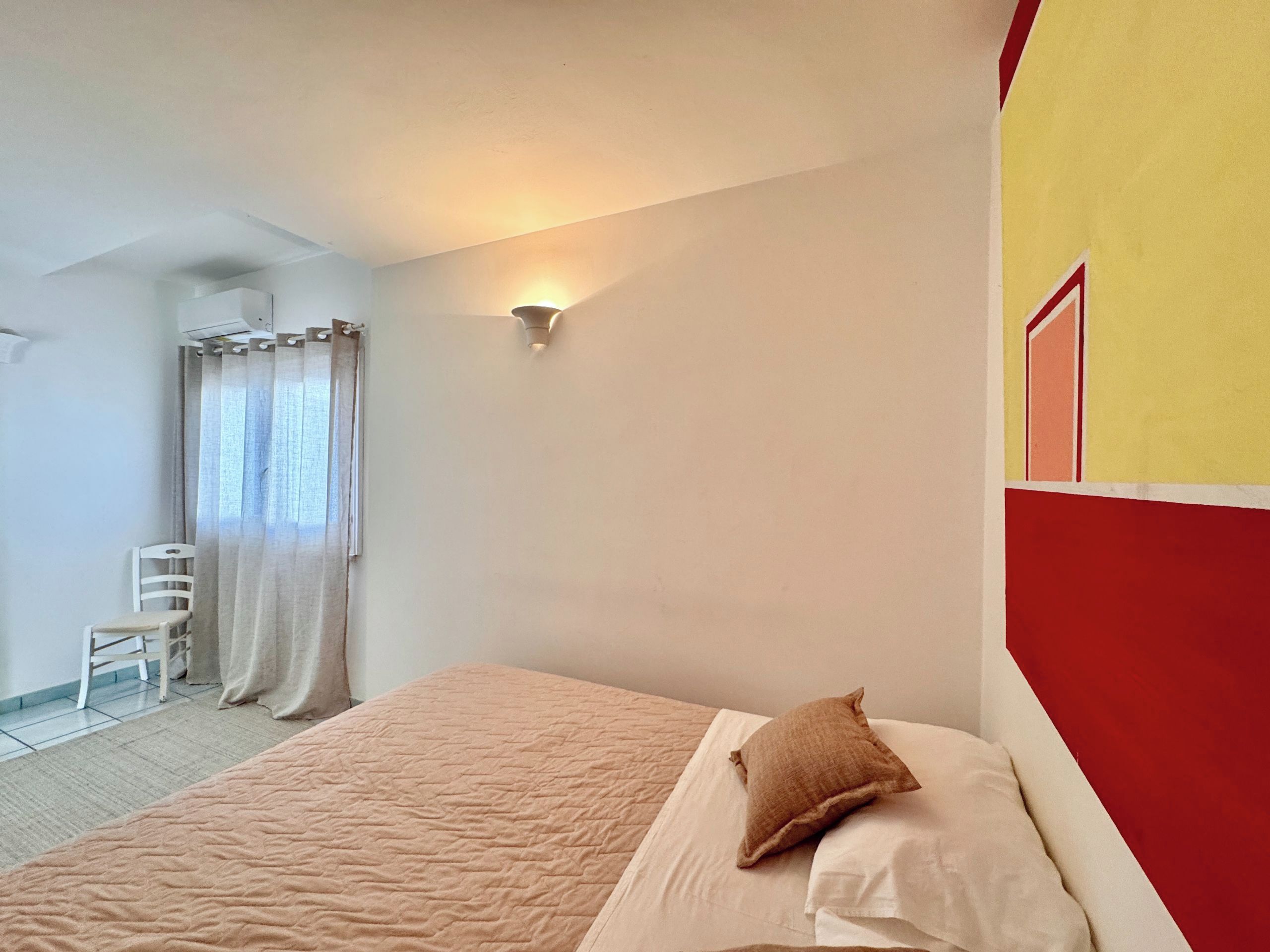 A bedroom with a bed, a chair, a window with curtains, and a wall-mounted light fixture.