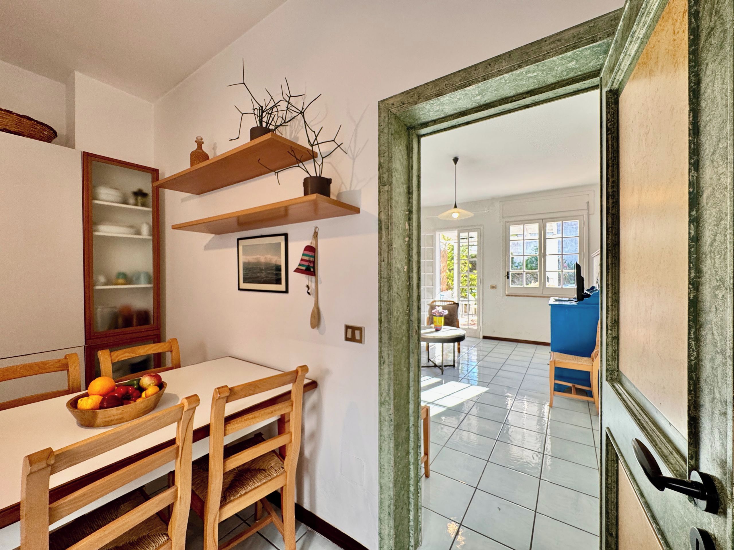 A dining area with a wooden table, chairs, and a fruit bowl. There is a wooden shelf above the table with decorative items. A door leads to another room with a blue chair and a table.