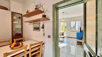 A dining area with a wooden table, chairs, and a fruit bowl. There is a wooden shelf above the table with decorative items. A door leads to another room with a blue chair and a table.