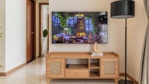 A living room with a flat-screen TV mounted on the wall, a wooden TV stand with a remote control, and a floor lamp. - Chao Nest Vinhomes Central Apartment (Ho Chi Minh City)
