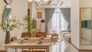 A modern living room with a sofa, dining table, and chairs. There is a large window with curtains, a ceiling fan, and a clock on the wall. - Chao Nest Vinhomes Central Apartment (Ho Chi Minh City)