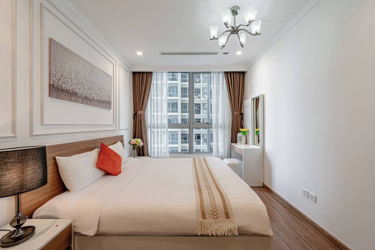 Une chambre d’hôtel avec un grand lit, une table de chevet, une lampe, une fenêtre avec des rideaux, un lustre et une œuvre d’art accrochée au mur.
