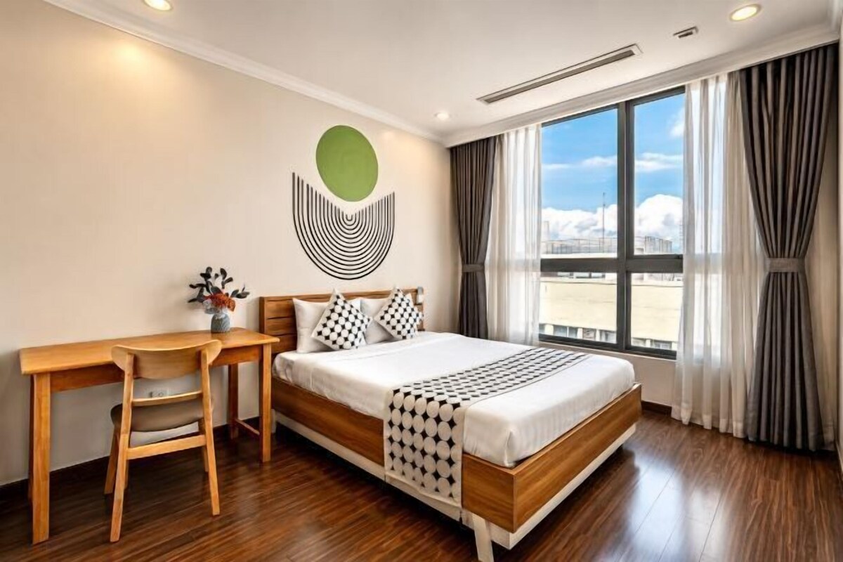 A hotel room with a large bed, a desk, a chair, and a view of the cityscape through the window.