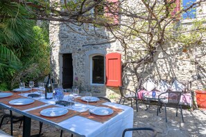 A stone building with a red shutter, a table set for dining with plates, glasses, and a bottle, and outdoor chairs.