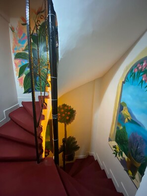 A staircase with red steps and a handrail, leading to a landing with a wall-mounted painting of tropical foliage.