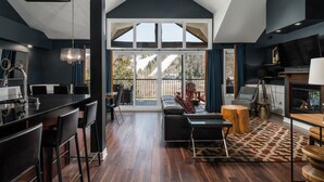 A spacious living area with a large sliding glass door leading to a deck, a fireplace, and a modern kitchen with a black countertop and bar stools.
