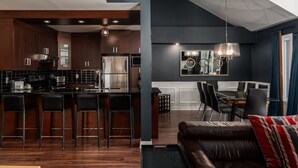 A modern kitchen with dark wood cabinets, stainless steel appliances, and a bar area. There is a dining area with a glass table and chairs, and a living room with a leather sofa and a large window.