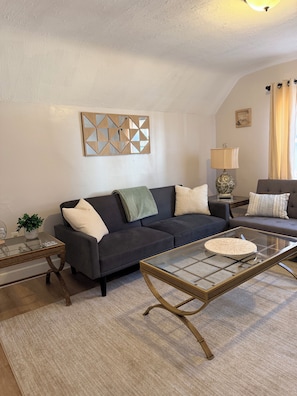 Una sala de estar con un sofá oscuro, una mesa de centro de vidrio, una lámpara y un espejo decorativo en la pared.