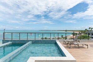 A rooftop pool with clear blue water, a view of the ocean, and a few lounge chairs. - Flat PiscinaPriv 1Quarto PortodeGalinhas (Ipojuca)