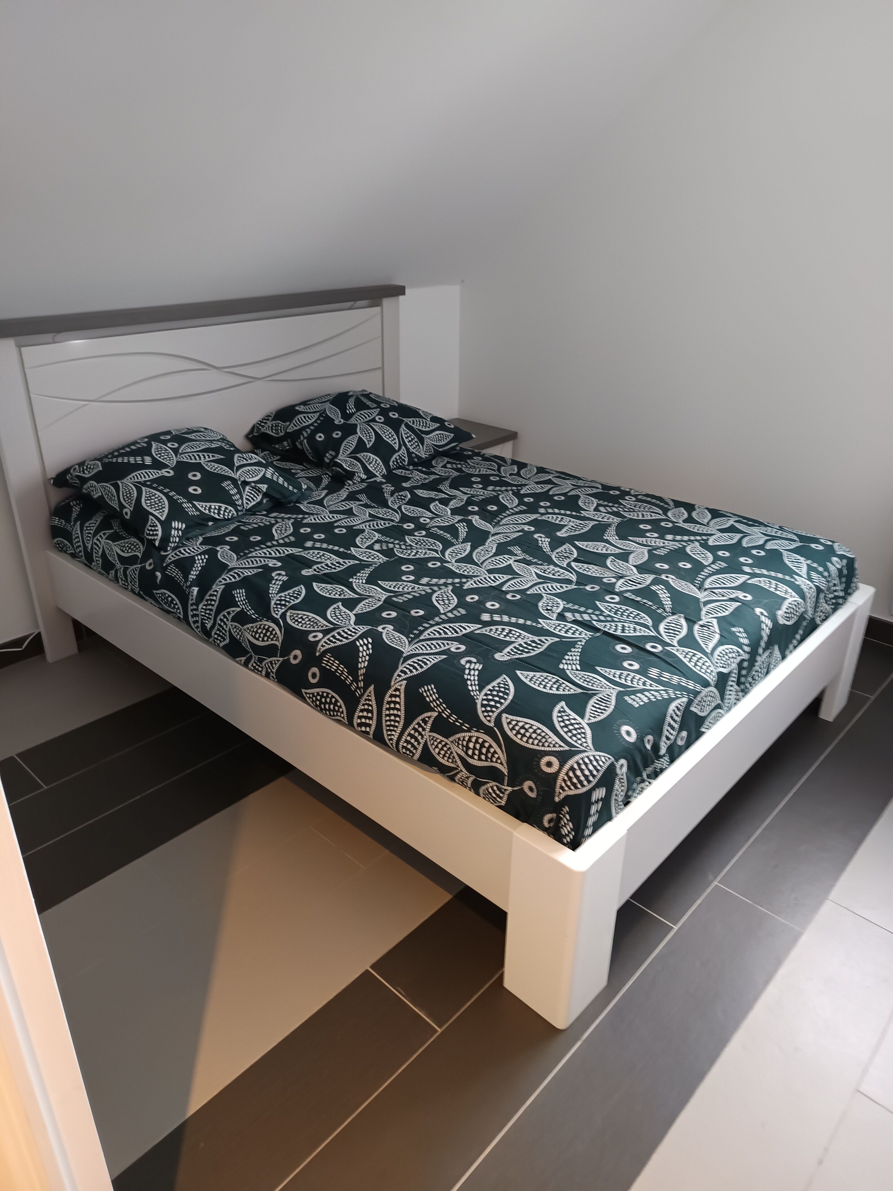 Un lit avec un cadre blanc et une couvre-lit vert foncé ornée d’un motif de feuilles.