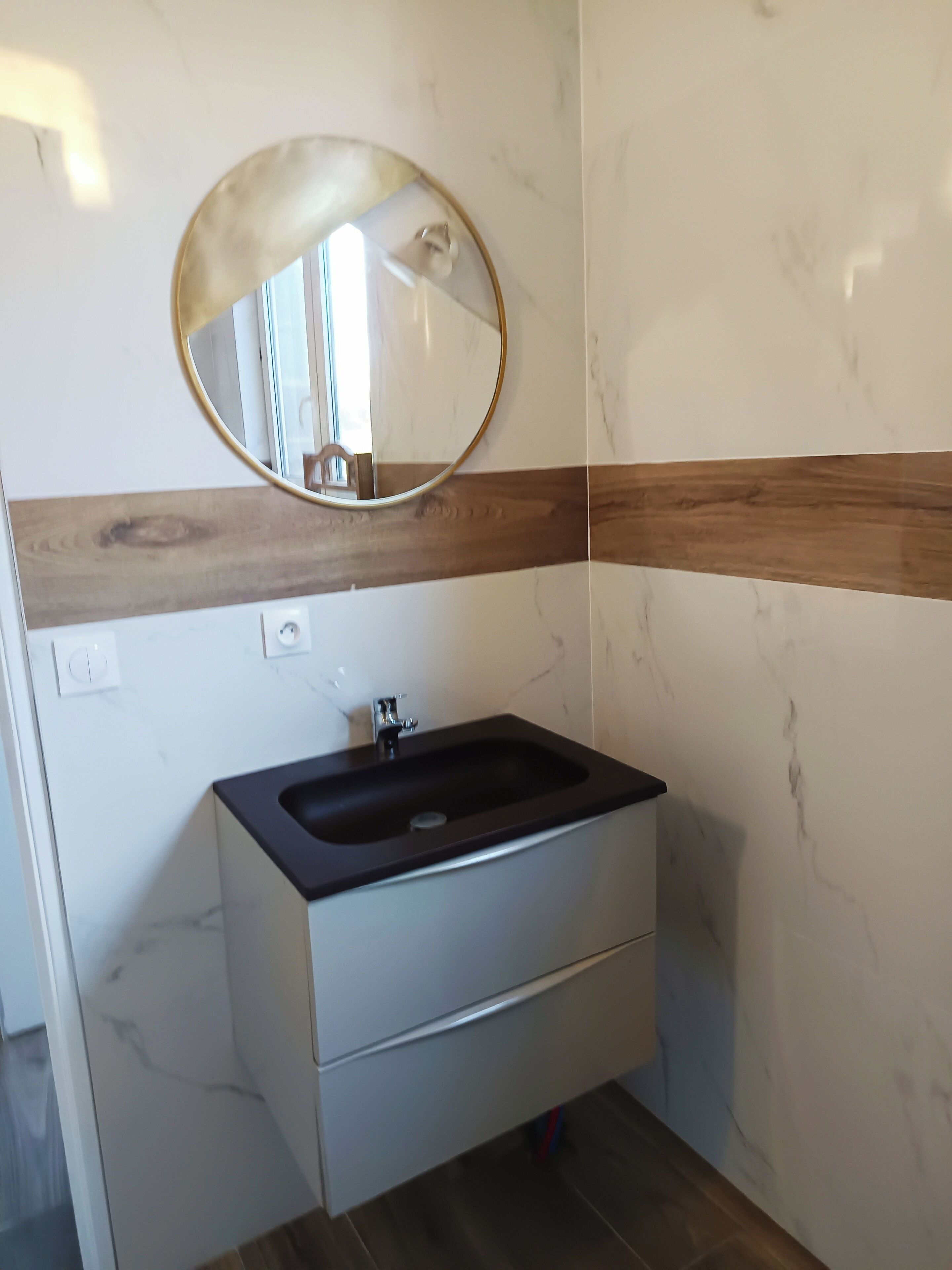 Une salle de bain avec un mur en marbre, une bande décorative en bois, un lavabo noir et un miroir.