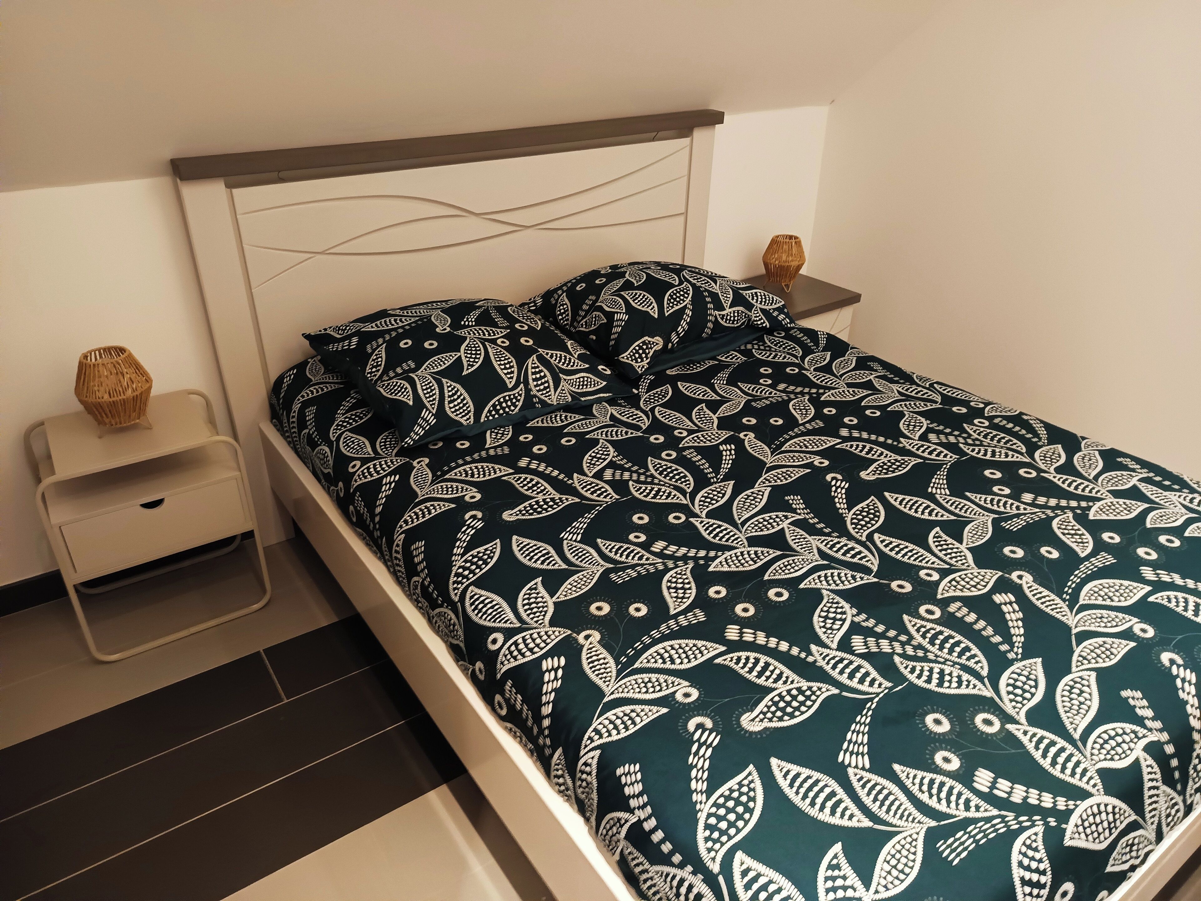 Een netjes opgemaakt bed met een donkere, gestreepte sprei en kussens, een nachtkastje met een geweven lamp en een ladekast.