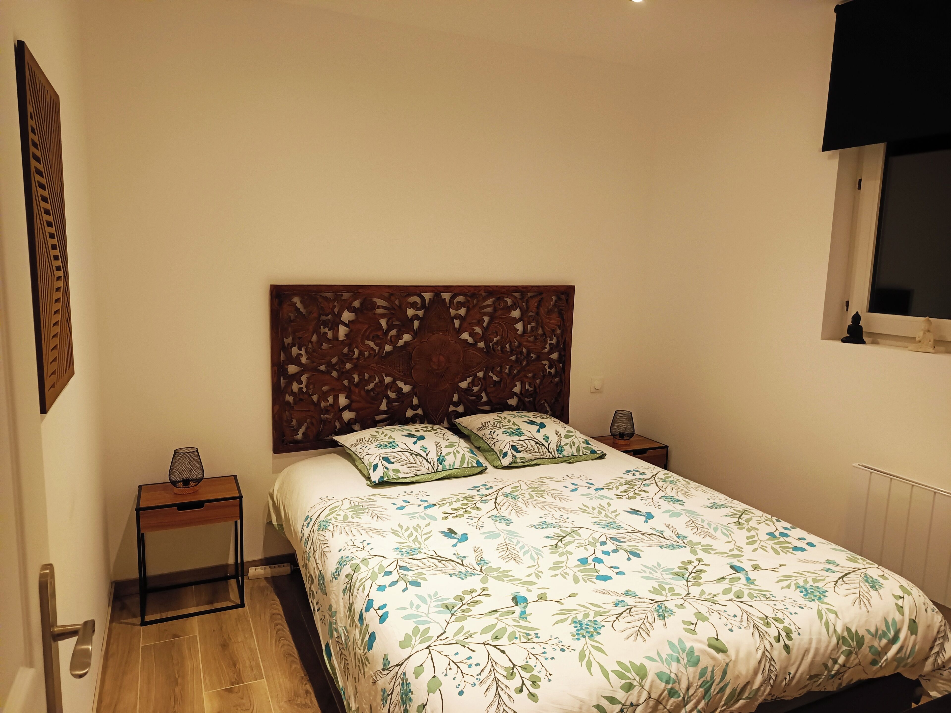 A bedroom with a wooden headboard, a floral bedspread, two nightstands with lamps, and a window with a view.