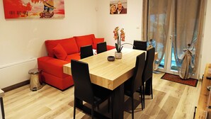 A modern living room with a red sofa, a wooden dining table, and black chairs. There is a small trash bin and a potted plant on the table.