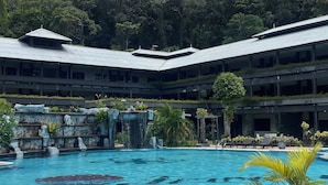 Une piscine dotée d’une plage carrelée, entourée de verdure, et un immeuble à plusieurs niveaux avec un toit plat.