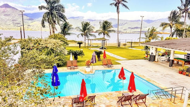 A pool area with lounge chairs and umbrellas, surrounded by palm trees and a view of mountains. - VELINN Hotel Guarda Mor (São Sebastião)