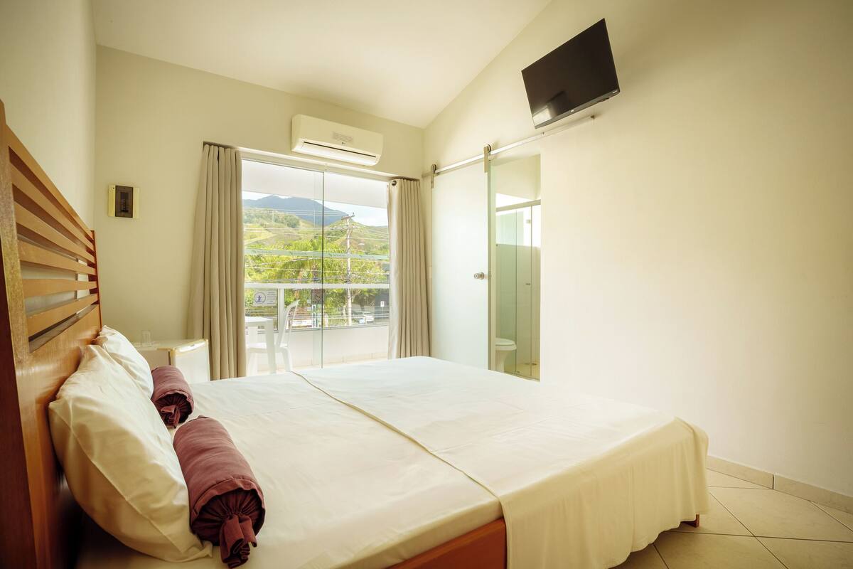 A hotel room with a bed, a TV, an air conditioning unit, and a view of greenery.
