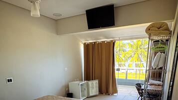 Habitación de hotel con cama, ventilador de techo, televisor montado en la pared, refrigerador pequeño, ventana con cortinas y balcón con una silla y estante con toallas.