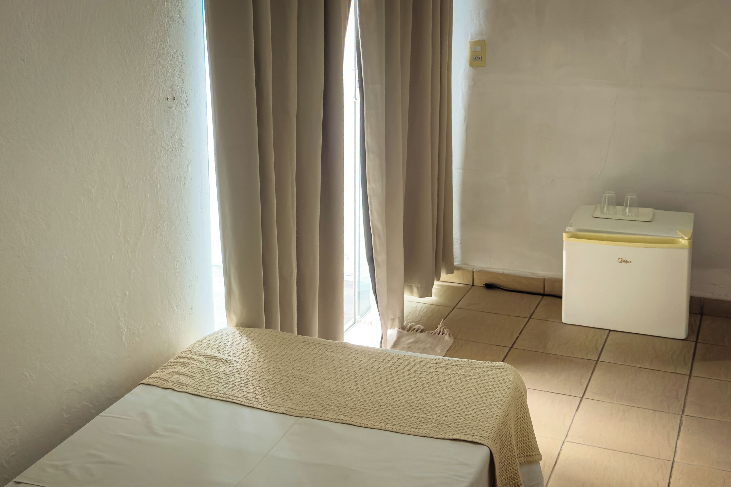 Una habitación con una cama, una mesita de noche pequeña, una lámpara y una ventana con cortinas.